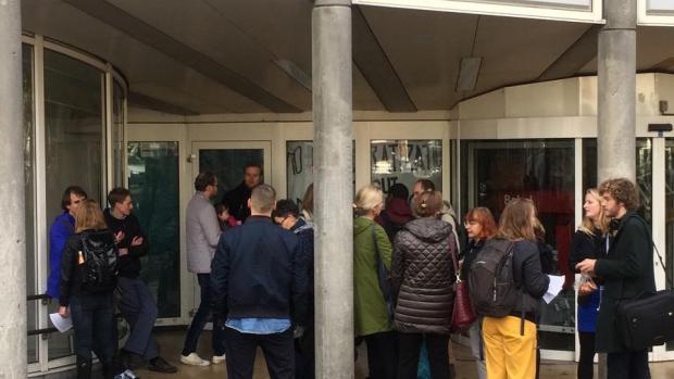 Colleges naar buiten verplaatst; docenten mogen spullen weghalen
Julia Prummel (21, europese studies) en Tesse Baardman (20, literary and cultural analysis) warmen hun handen aan hun thermosfles meegebrachte koffie op een bankje aan het Singel. Met een handvol studenten kregen ze het college Arabisch buiten op de gracht. &lsquo;Toen we het PCH niet in konden, verplaatste onze docent het college naar buiten. Ze wilde toch graag dat het doorging&rsquo;, zegt Julia. Al was het in het begin wel een beetje koud.&rsquo;
&nbsp;
Aan de achterkant van het PC Hoofthuis staat inmiddels een rij van zo&rsquo;n zes docenten die &eacute;&eacute;n voor &eacute;&eacute;n naar binnen mogen om spullen te halen. Vrolijk overleggen ze wie eerst mag. &lsquo;De deur moet zo kort mogelijk open&rsquo;, zegt een van de actie-woordvoerders. Als de deur opengaat, zijn vier jongens te zien, volledig in zwart, met bivakmutsen, sjaals en coltruien opgetrokken tot over de mond. Onder escorte wordt &eacute;&eacute;n opgewekte docent weer naar buiten gelaten.
