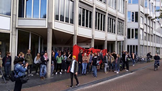 De studenten roepen nu verschillende leuzen en Geert ten Dam staat verderop
Aan de achterkant van het&nbsp;P.C. Hoofthuis wordt er tegen de studenten geroepen om in een cirkel op de grond te gaan zitten, arm in arm. Ze zitten nog niet, maar staan met een grote groep dicht op elkaar. 'Er zal gezongen worden!', wordt er geroepen. Dan&nbsp;galmen er verschillende leuzen over de grachten: 'Hoe laat is het? Solidariteit!', 'When education is under attack? Stand up, fight back!', 'He ho CVB, hier krijg je problemen mee!', 'No ifs, no buts, no education cuts!'. Verderop staat Geert ten Dam. Ze maakt nog geen aanstalten om naar de studenten toe te gaan.
&nbsp;